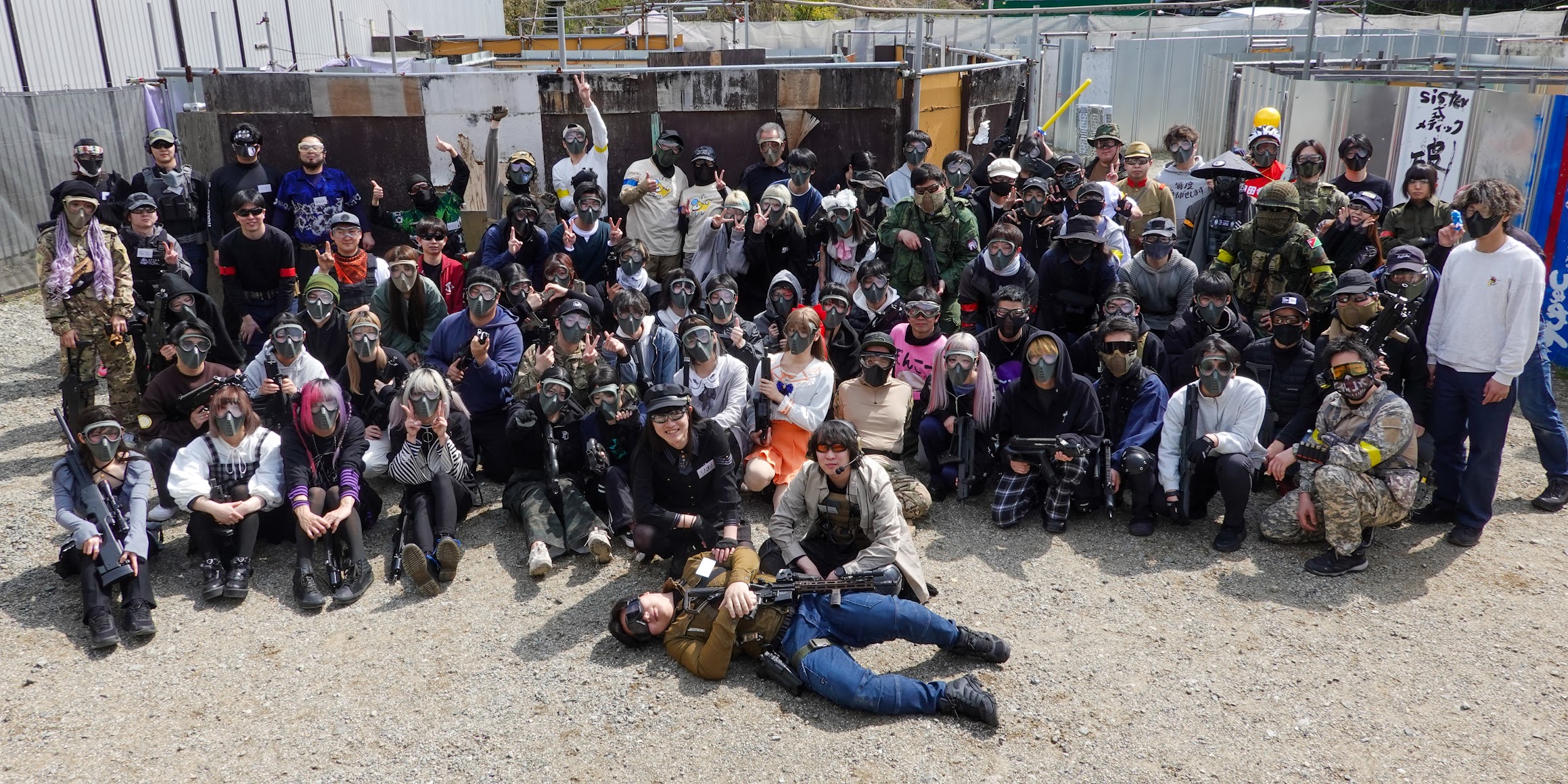 Large group of people wearing protective masks and tactical gear posing for a photo at an outdoor paintball/airsoft field.